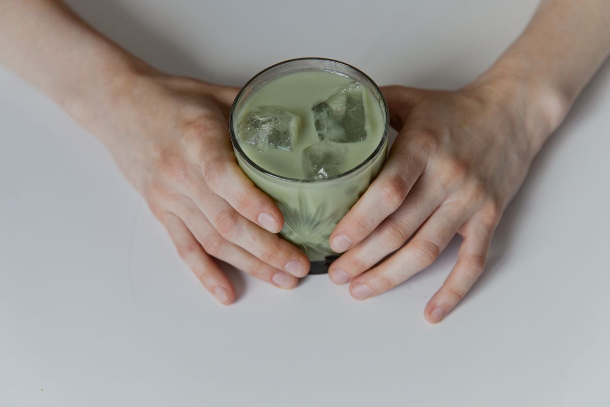 Hands holding a glass of iced matcha latte on a pale table