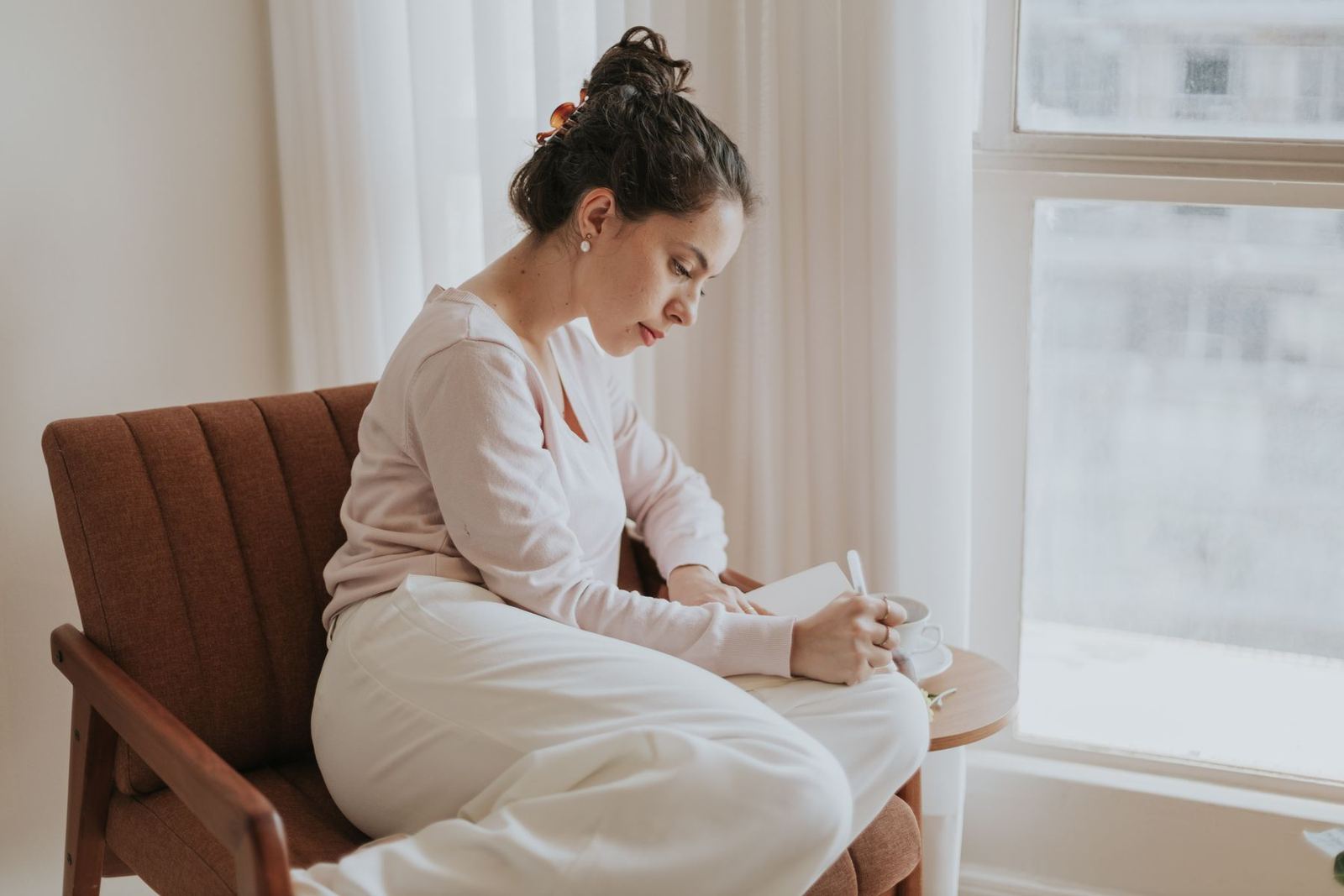 A woman in a cream sweater sits by a sunlit window, writing in a journal